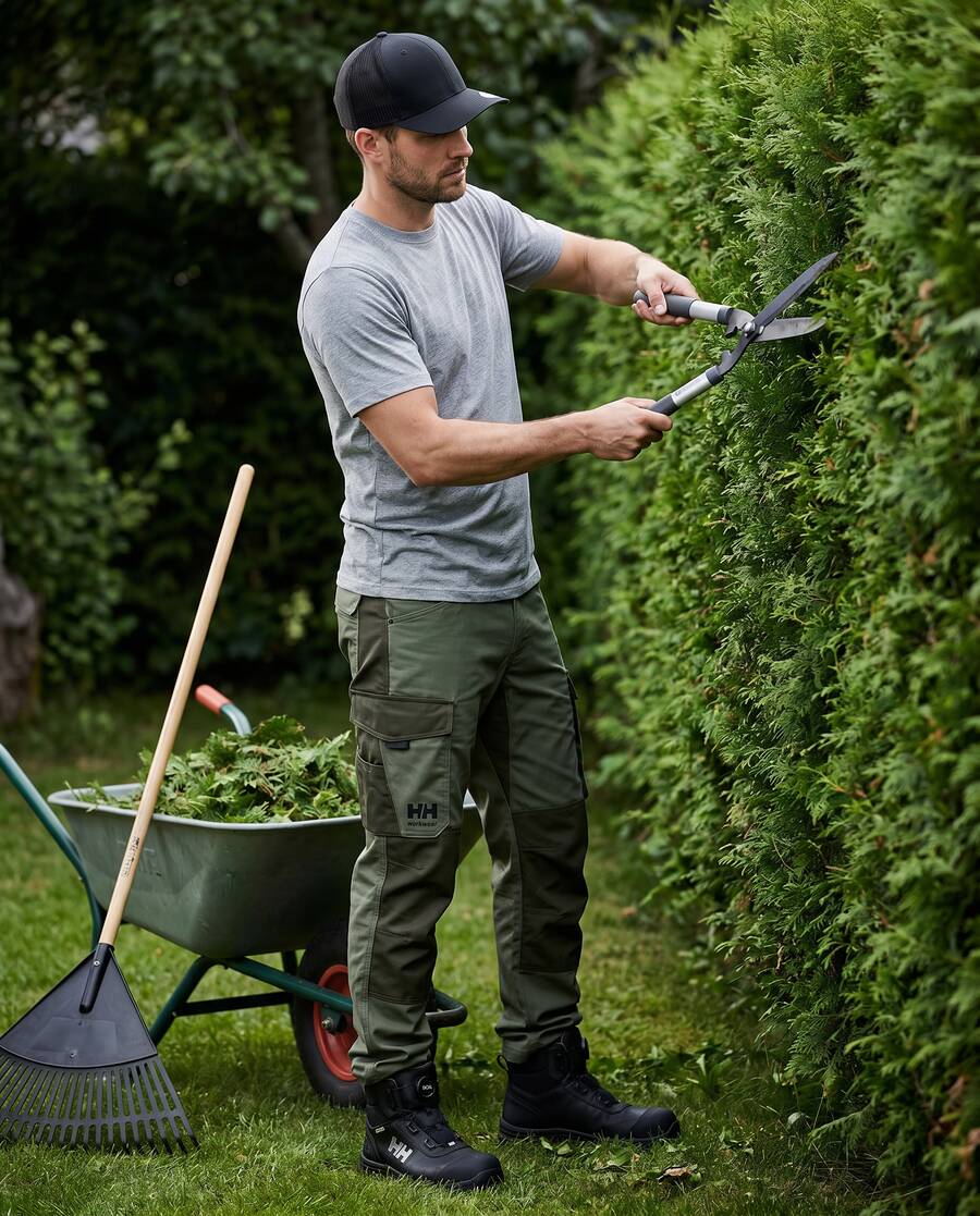 Gärtner schneidet mit einer Heckenschere eine Thuja-Hecke, Schubkarre mit Grünschnitt im Vordergrund