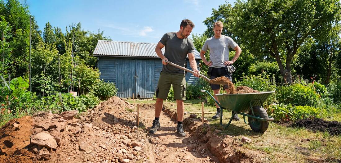 Zwei Männer schaufeln Erde in eine Schubkarre neben einem Gartenschuppen